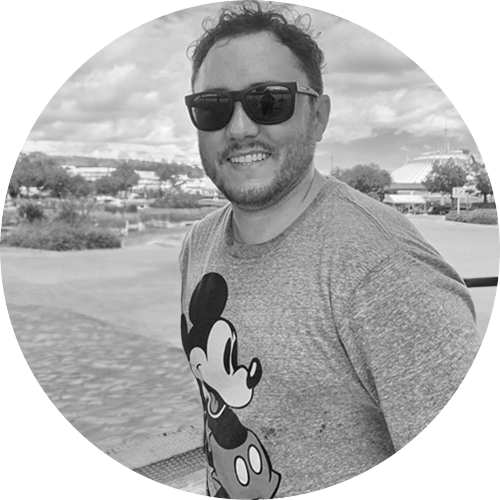 Smiling man wearing sunglasses and a Mickey Mouse t-shirt outdoors with trees and buildings in background.