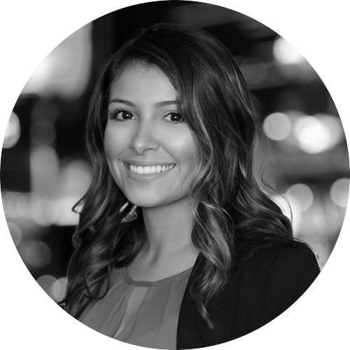 Smiling woman with wavy hair wearing a blazer against a blurred background in black and white.