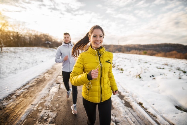 A man and woman jog in the snow, both wearing winter running gear and enjoying the cold weather exercise.