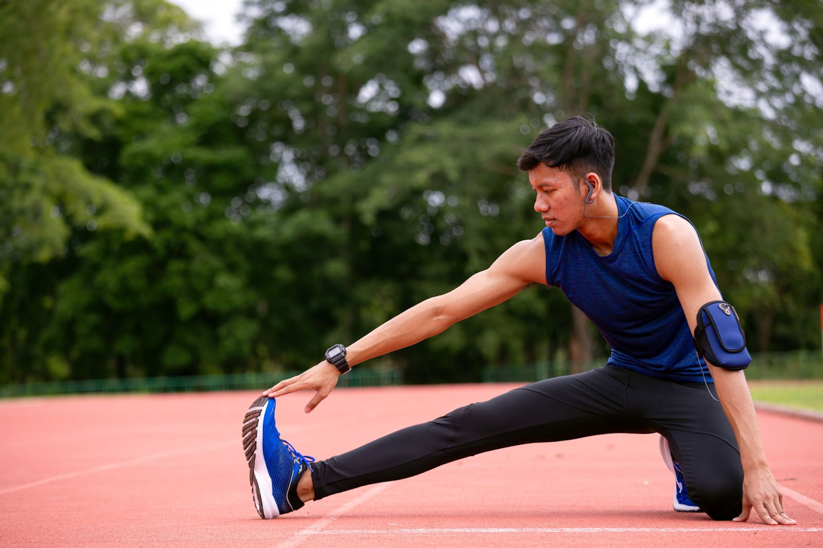 A young man in a blue sleeveless shirt performs a seated hamstring stretch on a red outdoor running track.