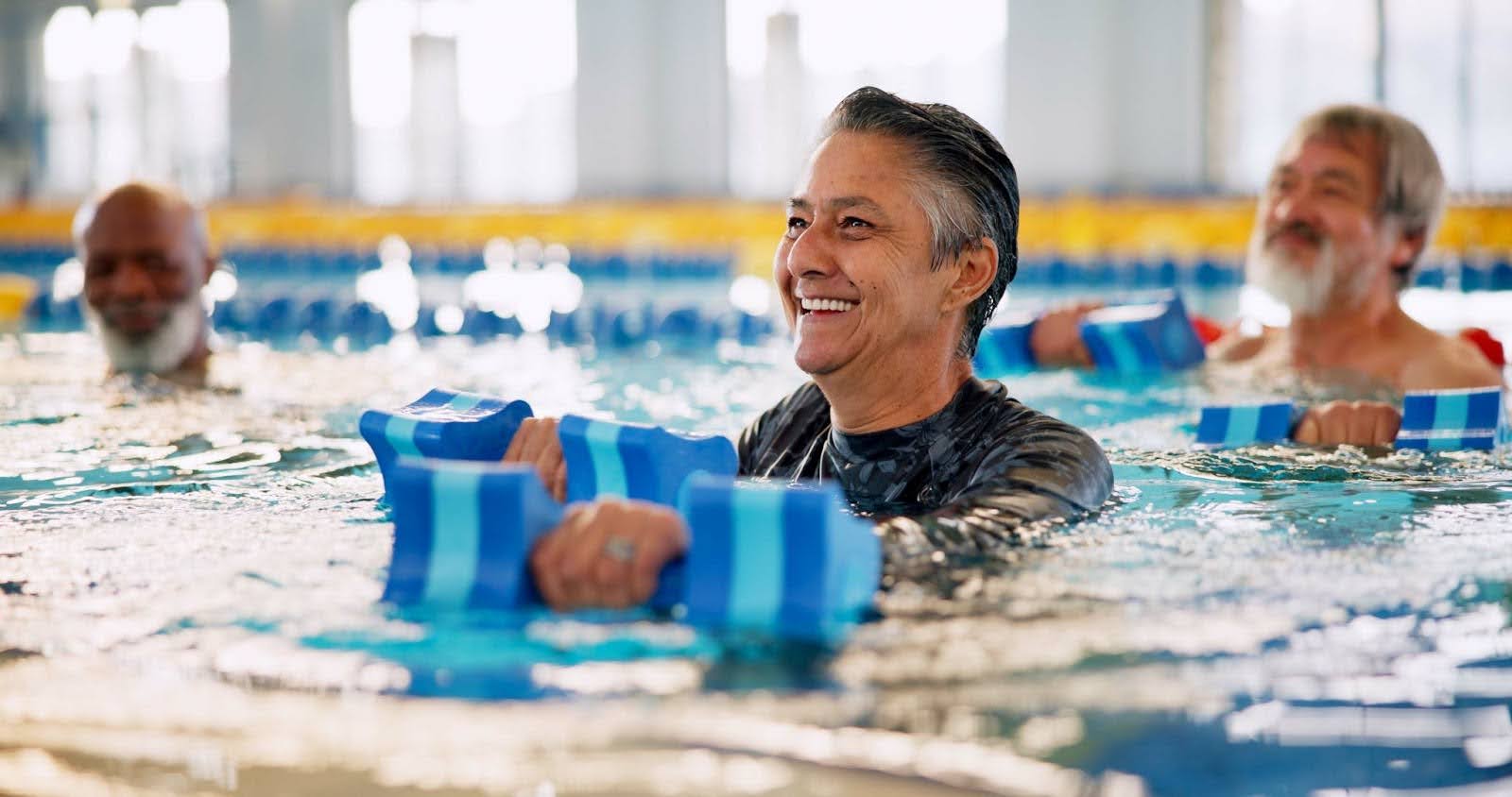 A group of senior citizens smiling while participating in an water aerobics class in a swimming pool, using blue foam dumbbells.