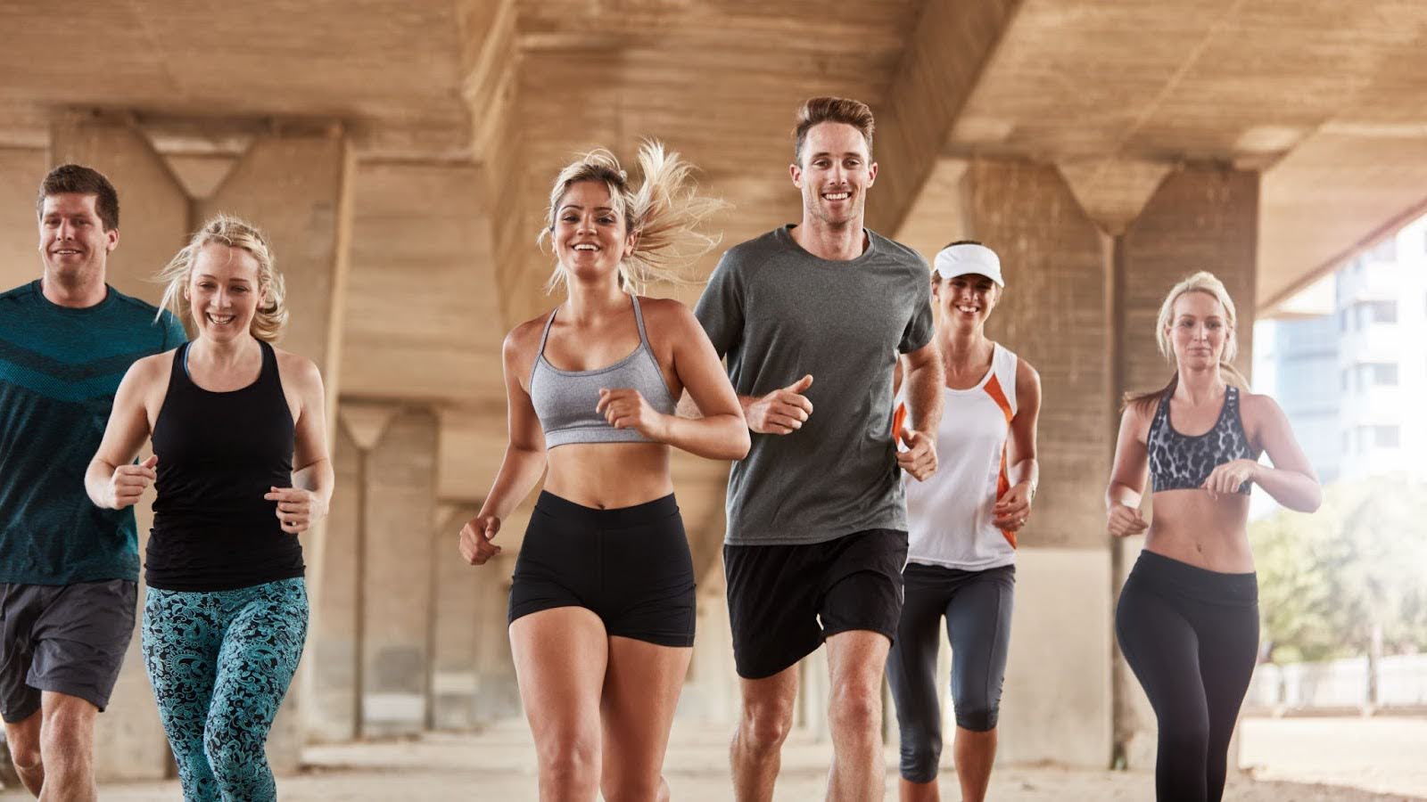 A group of smiling men and women in various athletic wear jogging together under a concrete urban bridge.