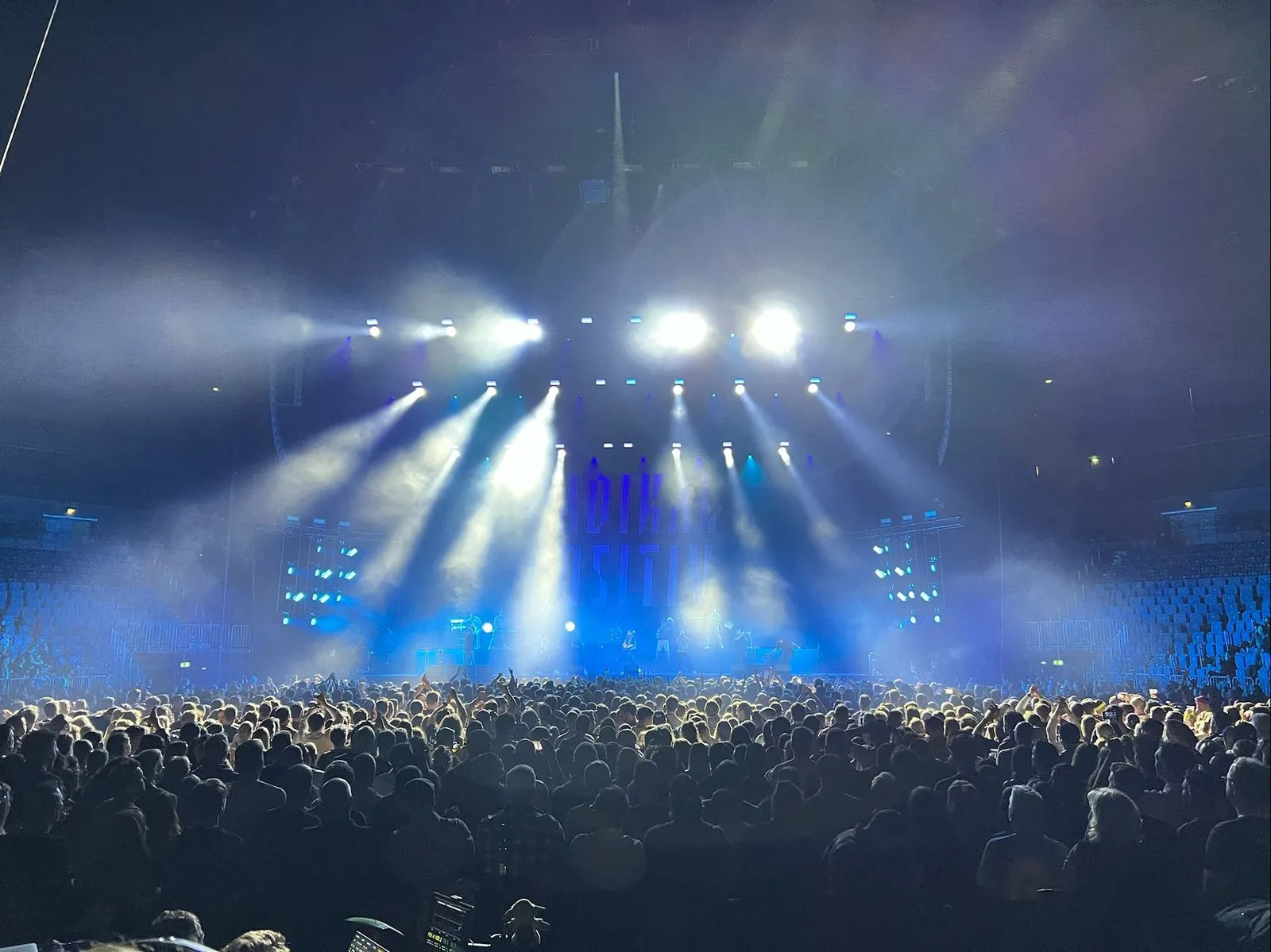 Ein konzert von Querbeat in der Kölner Lanxessarena. man sieht eine Bühne mit weißen lichtstrahlen, die ins publikum gerichtet sind. der Hintergrund ist in einem hellen blauton gehalten. Das Publikum jubelt der Band zu