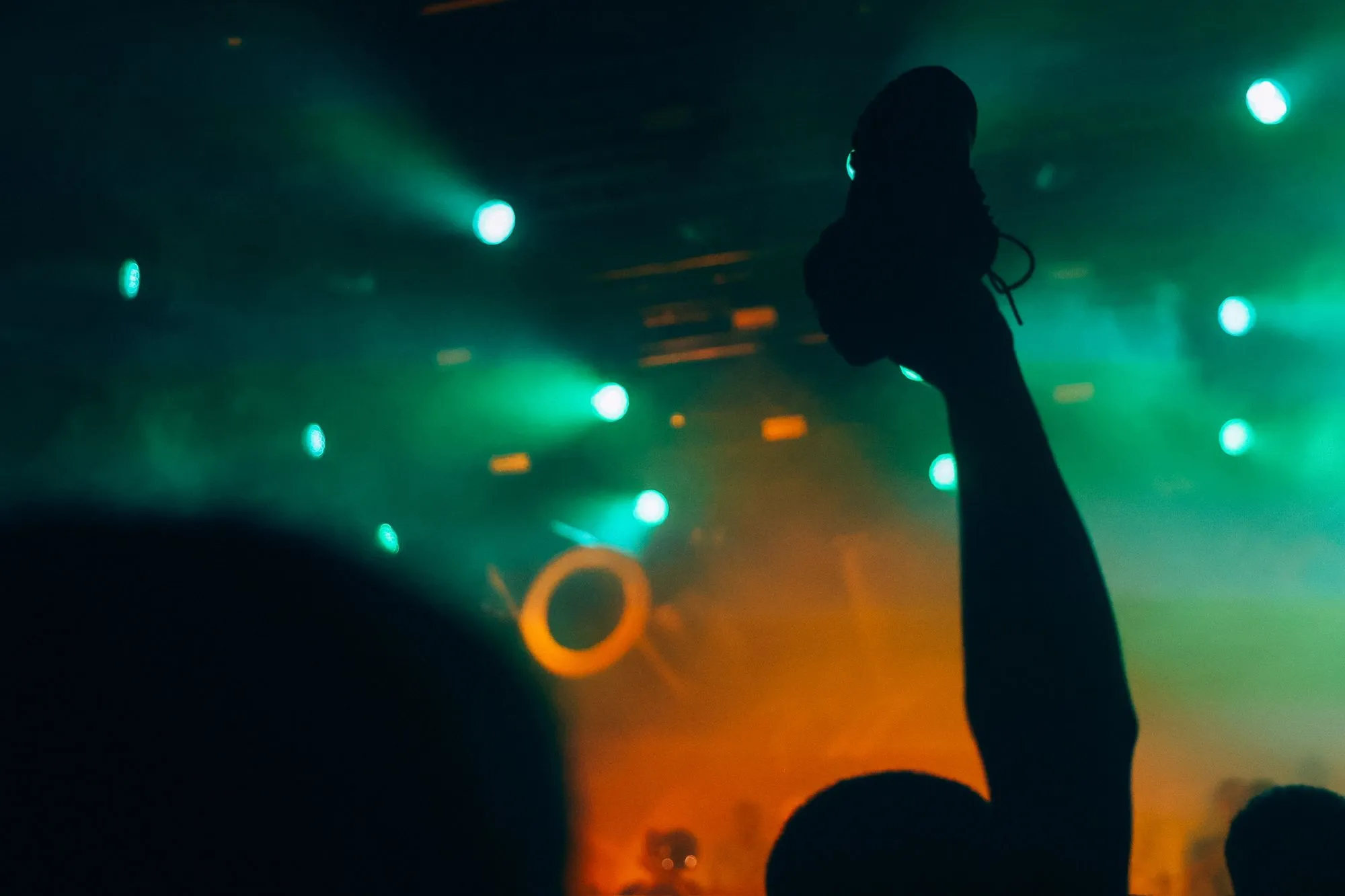 A person's arm is raised among a crowd at a concert, with colorful stage lights overhead.