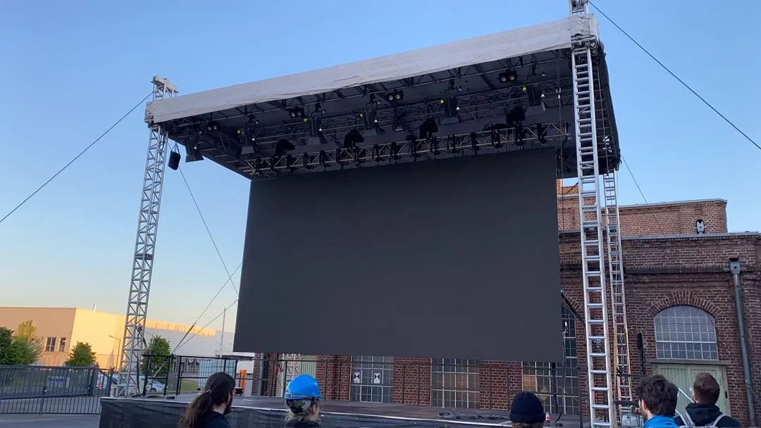 An outdoor stage with a large, blank screen and several people standing in front.