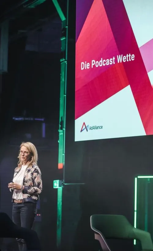 Eine Frau steht auf einer Bühne und gibt eine präsentation. Im Hintergrund zeigt die LED Wand den Titel:"Die Podcast Wette."