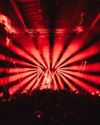Eine Menschenmenge steht vor einer Bühne auf der ein DJ untermalt mit Lichttechnik, performt. Die Lichtstrahlen sind orange