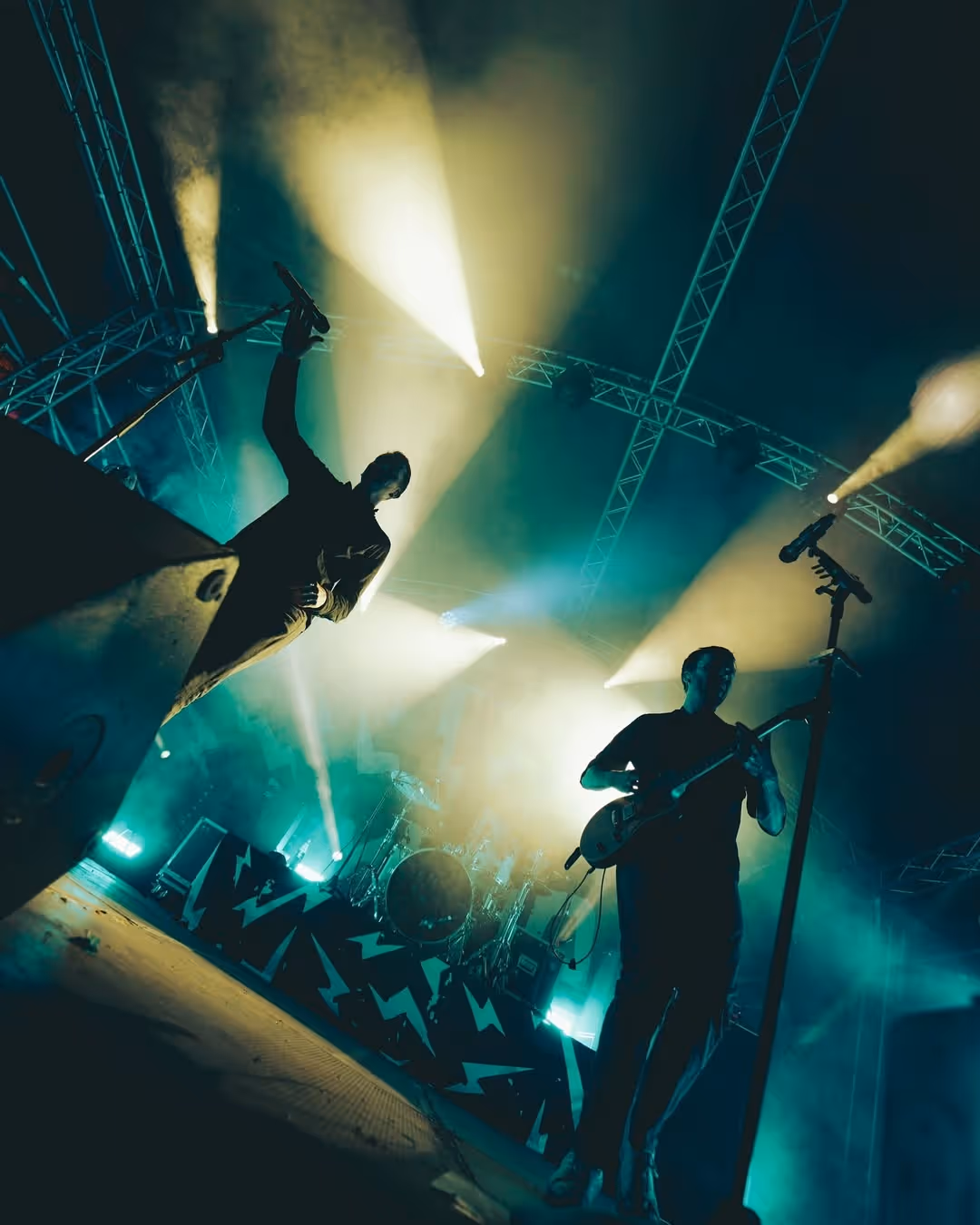 Zwei Musiker stehen auf einer Bühne und performen, energetisch mit Gitarre und Gesang und werden von Lichttechnik beleuchtet