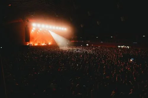 Eine große Menschenmenge steht vor einer Bühne und schaut ein Konzert. Die Blind Scheinwerfer sind eingeschaltet und orangenes Licht kommt von hinten. In der Mitte ist ein Spot, der in das Publikum geht.