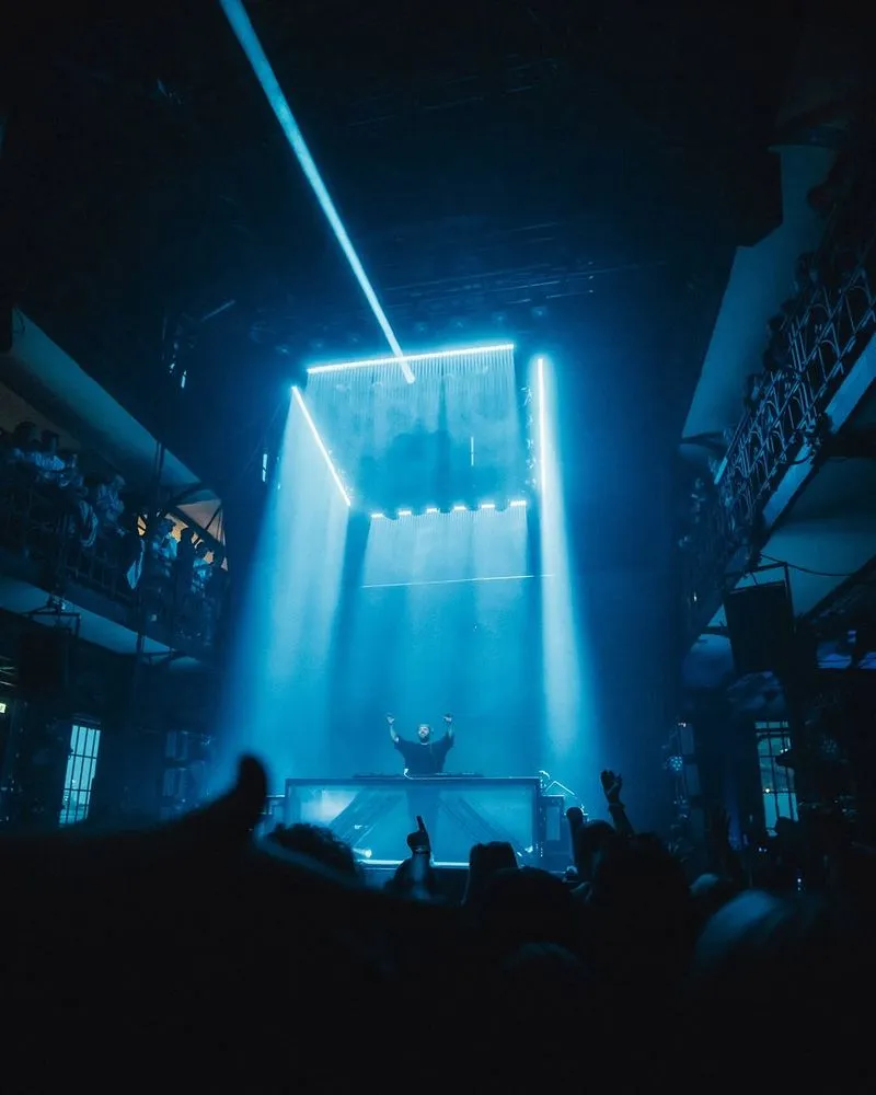 Ein DJ steht an seinem DJ Tisch, der mit Milchglas verkleidet ist. Über ihm ist ein Vorhang von blauen Licht zu erkennen, der in einem Quadrat angeordnet ist. Von oben ist Lichttechnik von photon zu sehen.