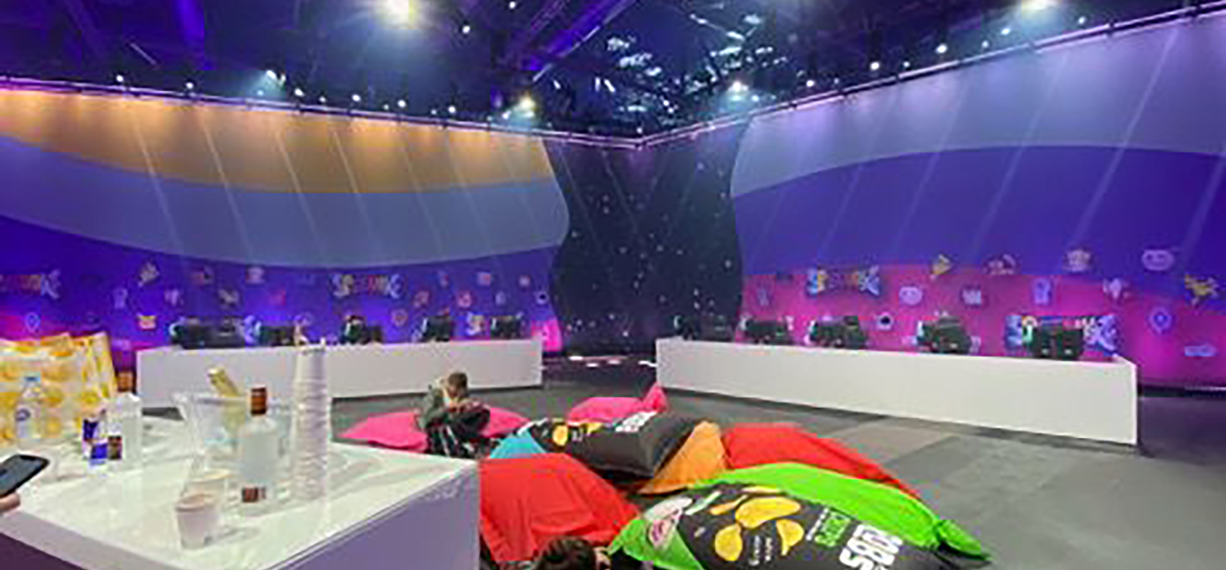 Man sieht eine Lange tafel aufgereit mit drehstühlen, an jedem Platz ist ein PC aufgebaut. von oben kommen weiße Lichtstrahlen und im Hintergrund ist ein Buntes Banner gespannt