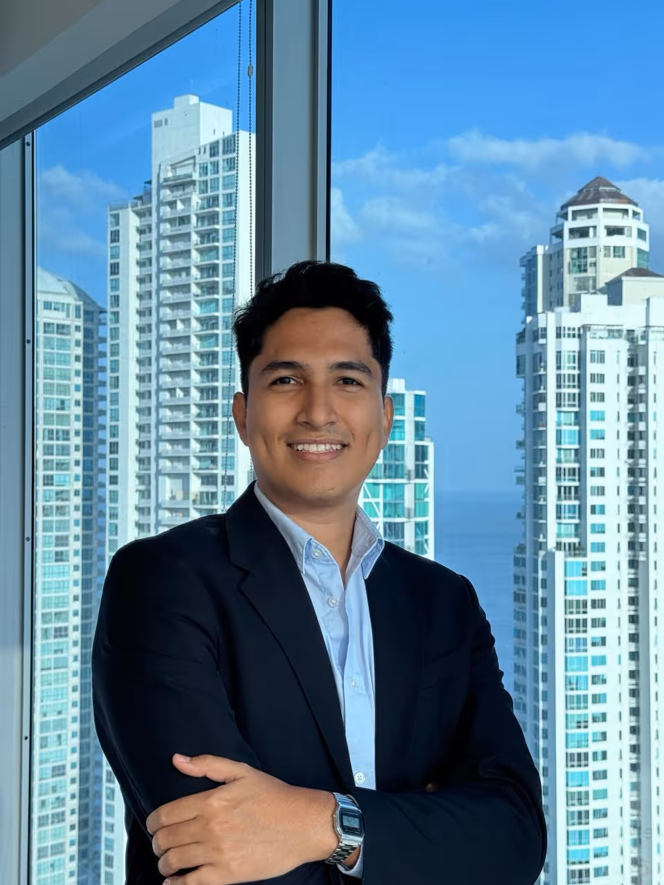 Smiling man in a black blazer and light blue shirt standing with arms crossed in front of a window with a cityscape of tall buildings and blue sky.