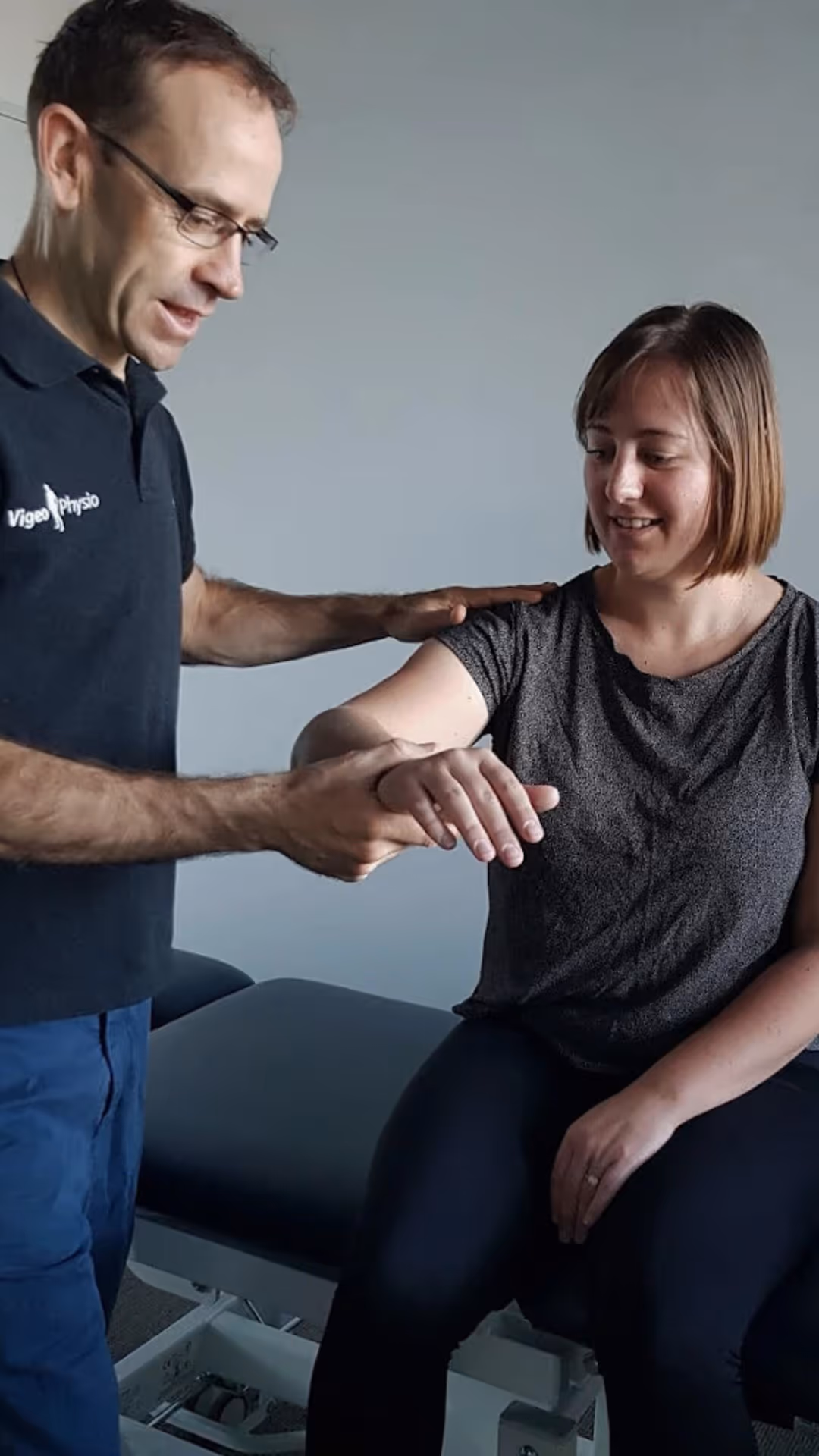 A physiotherapist (Dominic Brosnahan from Vigeo Physio) assisting a seated woman with arm movement during a therapy session.