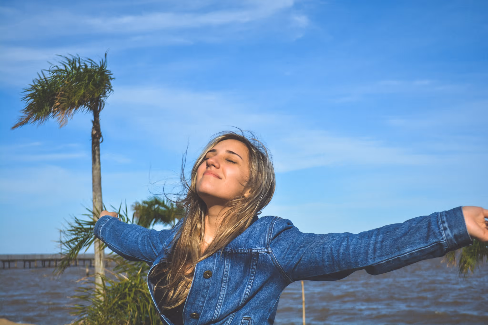 Young woman in a denim jacket standing by water with arms outstretched and eyes closed under blue sky.