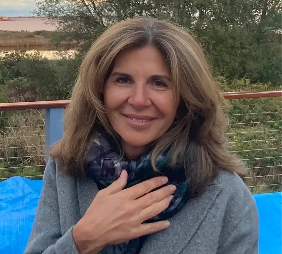A portrait CEO Stephanie Georges wearing a gray sweater and a dark patterned scarf, standing outdoors with greenery and a body of water in the background.