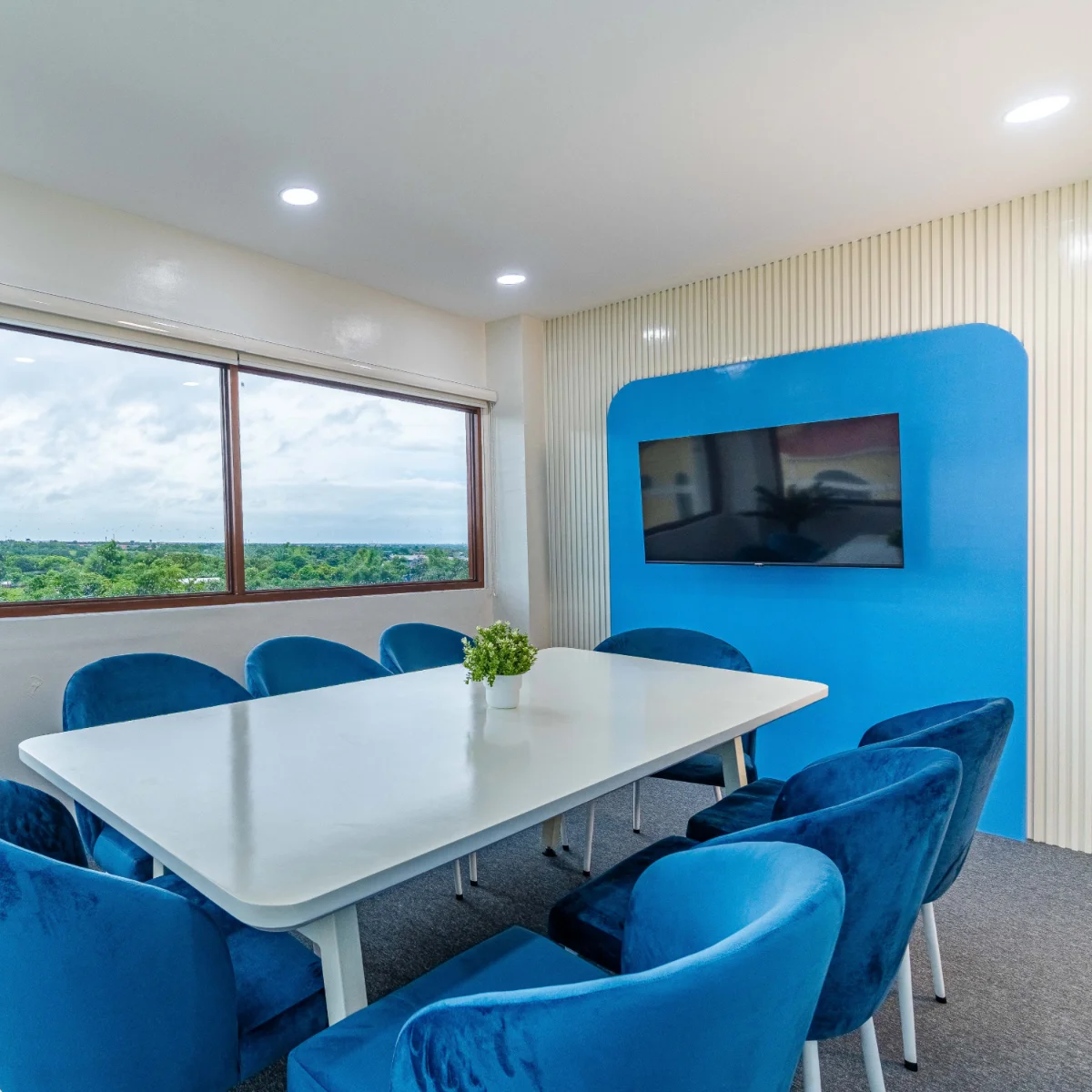 Bright conference room with a large white table surrounded by plush blue chairs, facing a wall-mounted flat screen on a bold blue accent wall. A wide window offers a scenic view of green landscape under a cloudy sky, adding natural light to the clean, modern space.