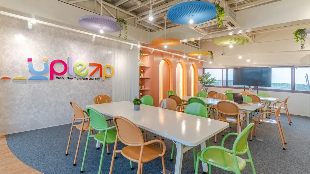 Colorful and modern coworking space with white tables, green and tan chairs, and circular ceiling panels in blue, orange, yellow, and purple. A vibrant sign on the wall reads "Upleap – Work. Play. Transform your day." and the room is accented with greenery and warm lighting.