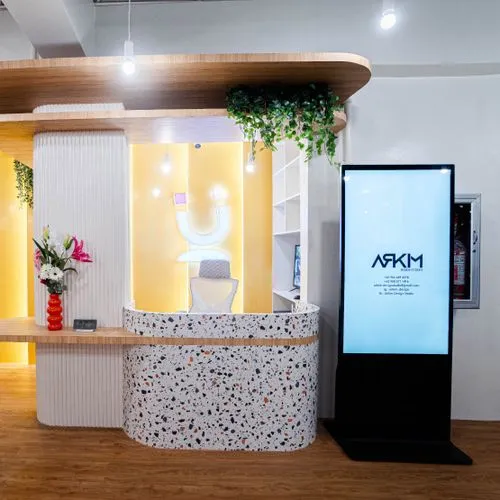 Modern reception area with a terrazzo-patterned front desk, a glowing yellow backdrop, and a backlit "U" logo above a white office chair. To the right, a digital signage display reads “ARKIM” and lists office details, while greenery and flowers add a welcoming touch.