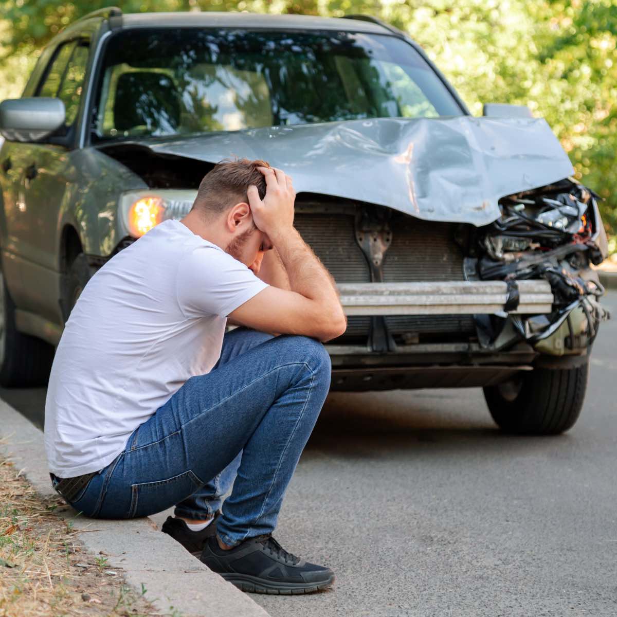 Man sitting on the roadside with his head in his hands beside a silver car with a heavily damaged front end.