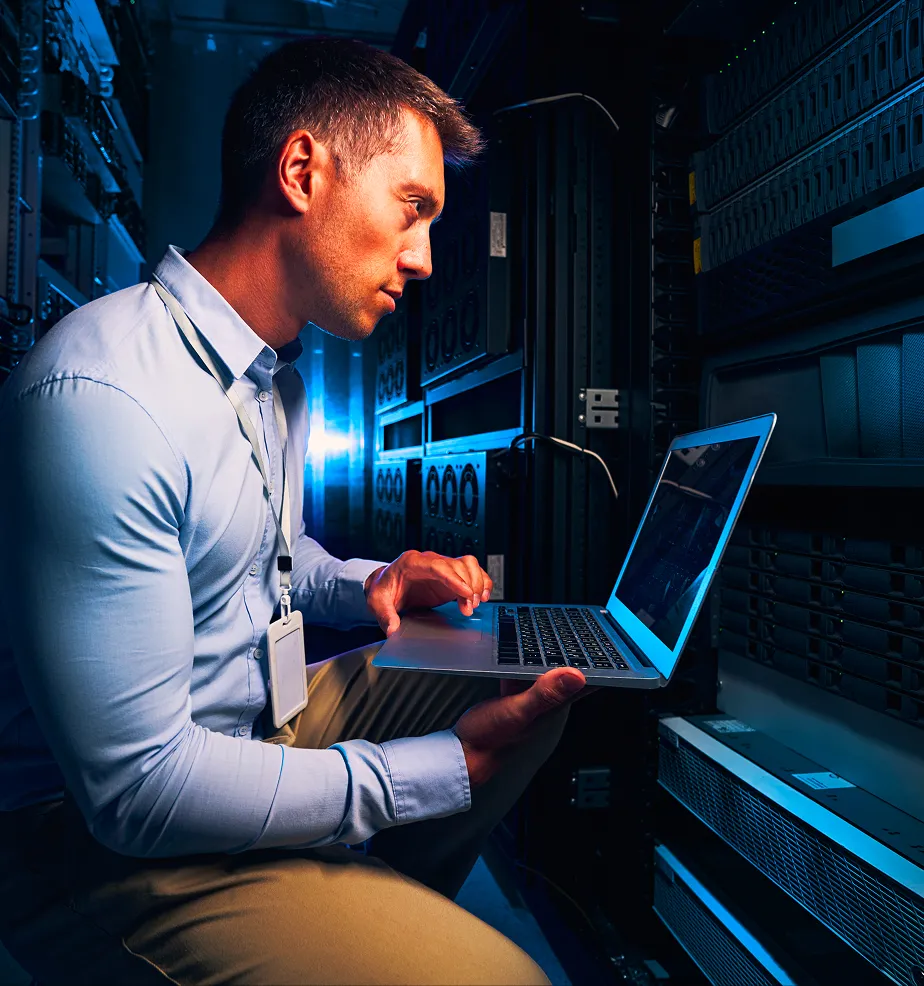 Man in a light blue shirt working on a laptop in a server room.