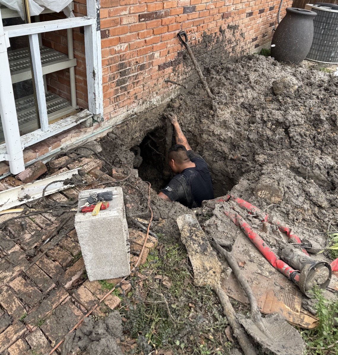 Worker repairing plumbing in a deep muddy hole next to a brick house.