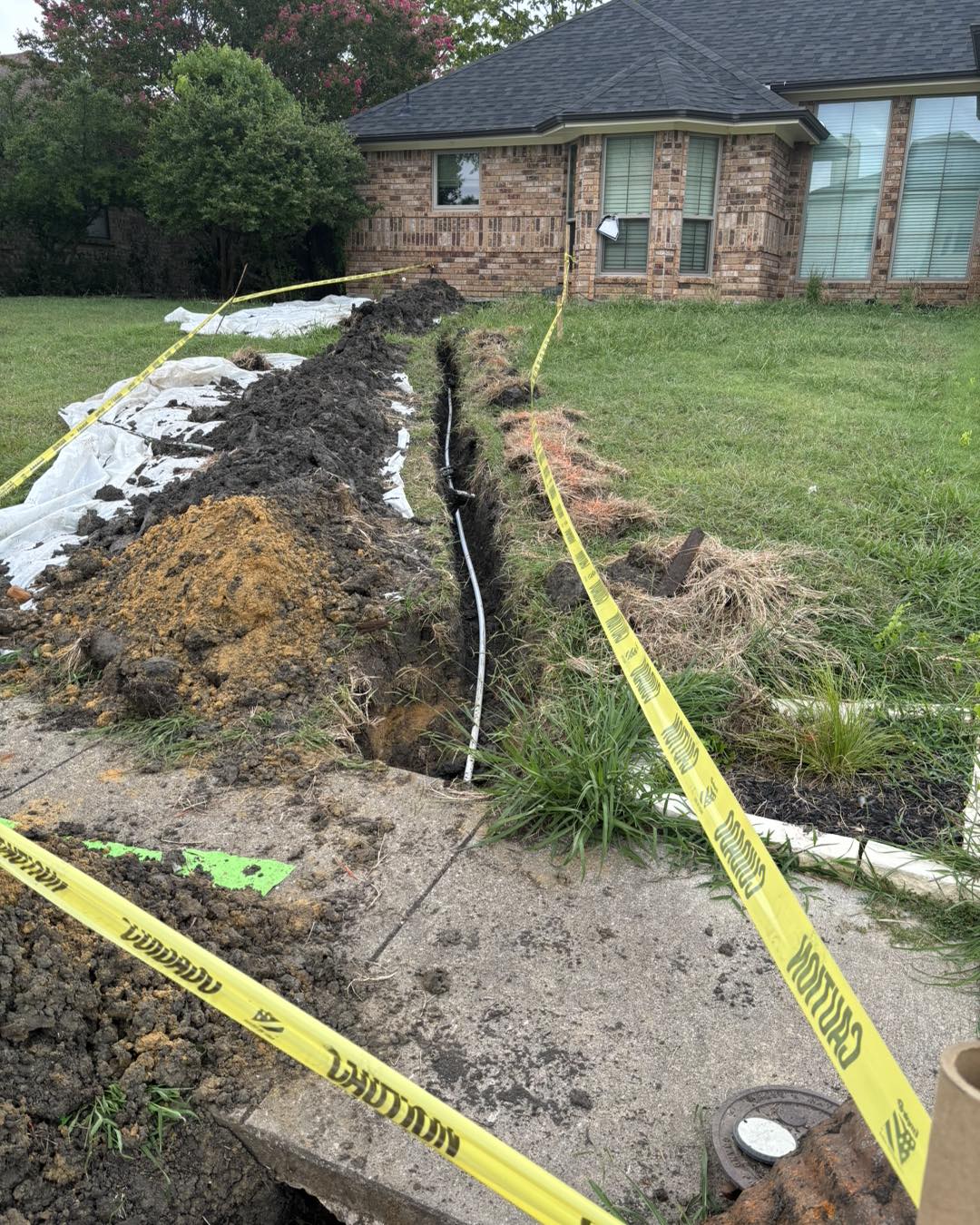 Long trench dug across a yard with caution tape marking a pipe repair.