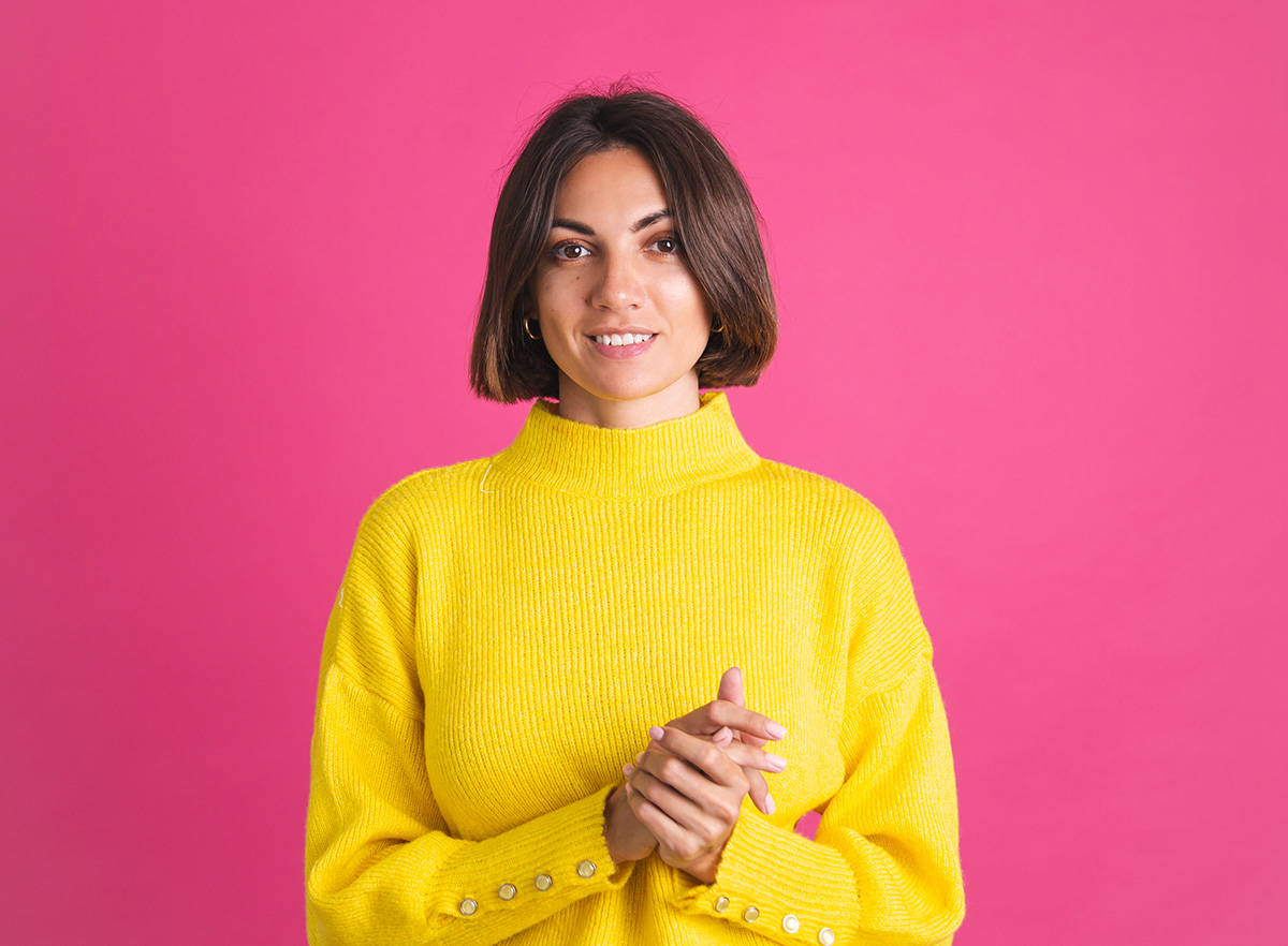 Woman with short brown hair wearing a bright yellow sweater smiling against a pink background.
