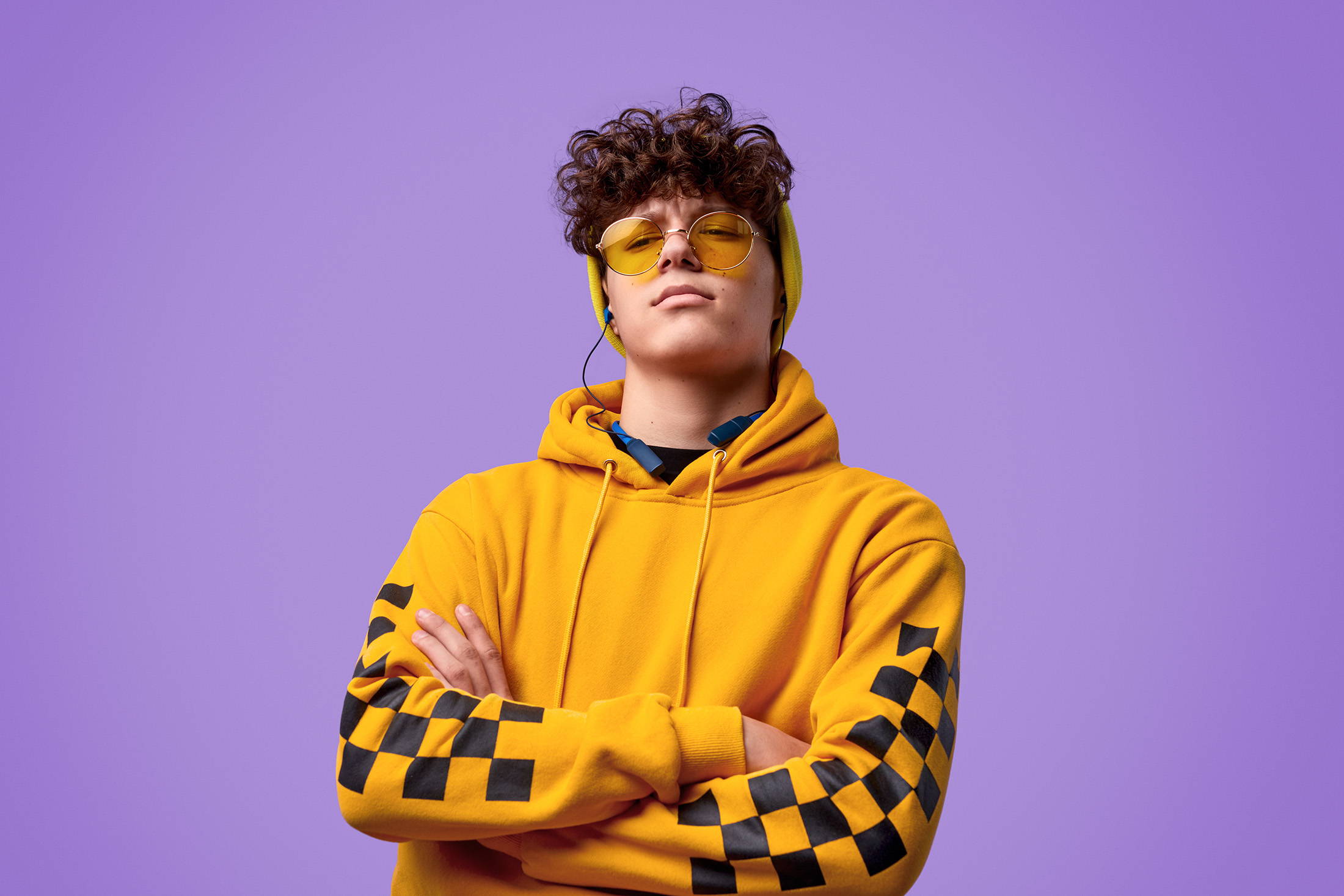 Confident teenager wearing a yellow hoodie with black checkered sleeves, yellow sunglasses, and a matching beanie, standing with arms crossed against a purple background.