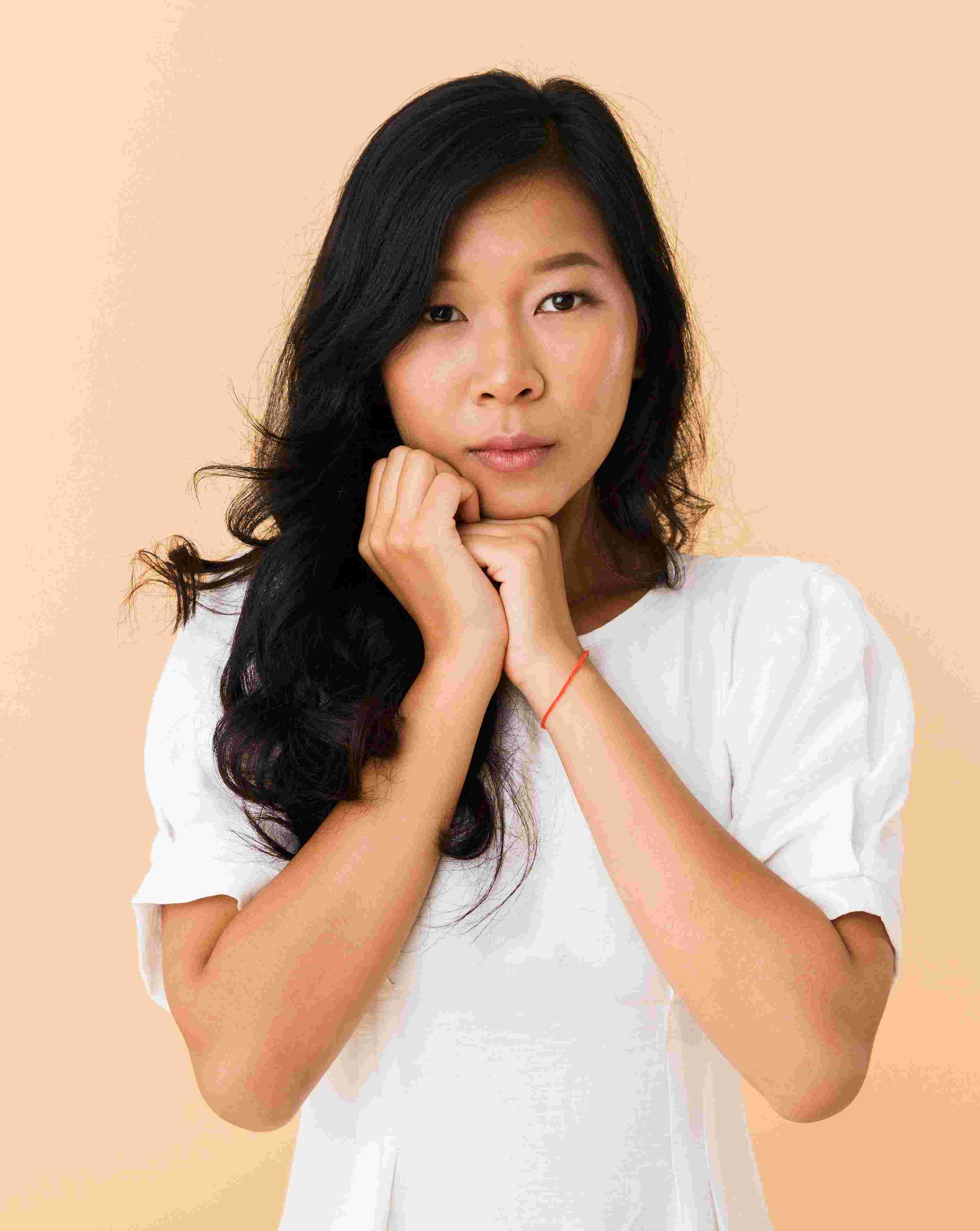 Portrait of a young woman with long black hair and a white shirt, resting her chin on her clasped hands against a beige background.