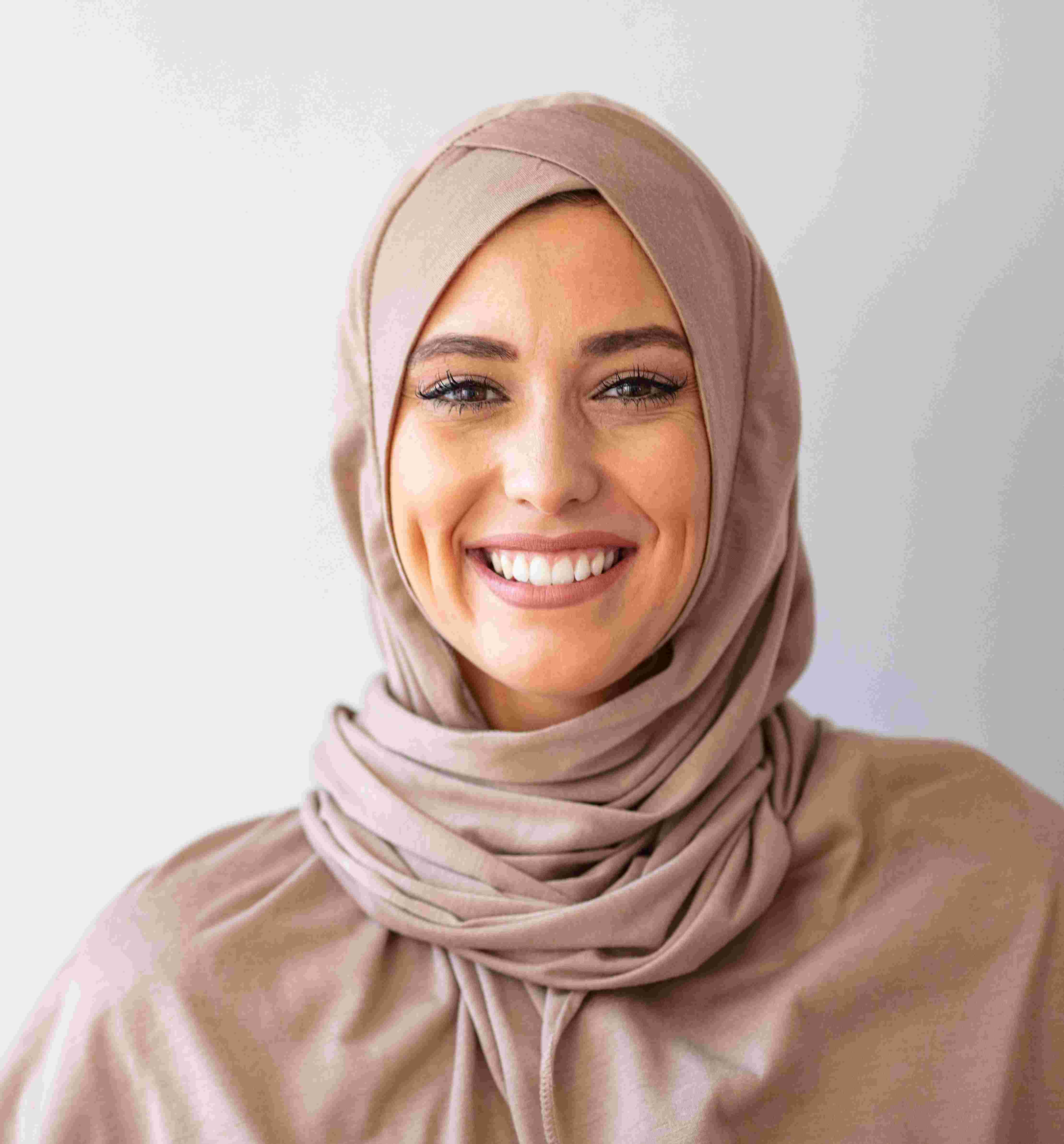 Smiling woman wearing a light brown hijab against a plain light background.