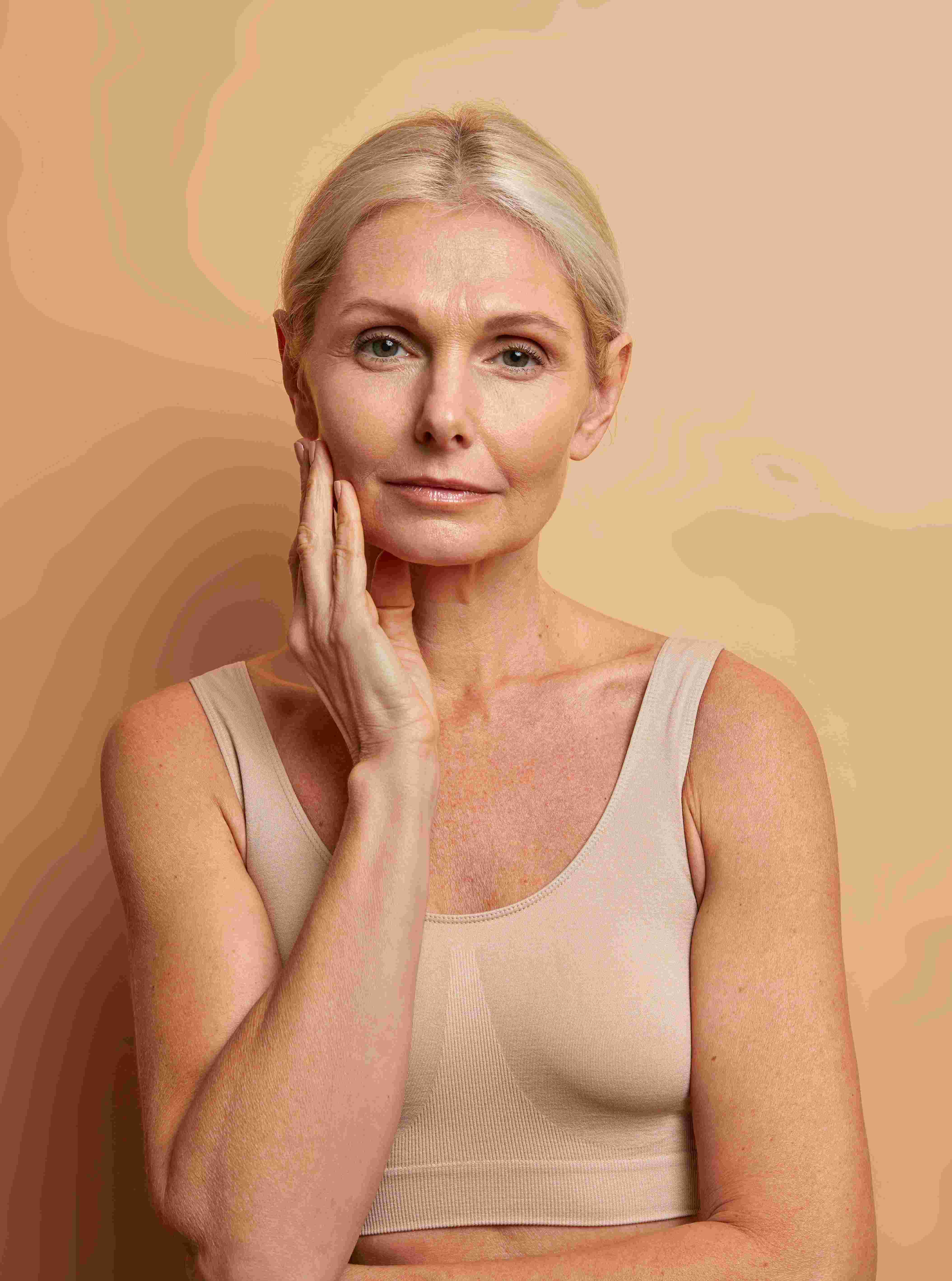 Mature woman with blonde hair touching her face, wearing a beige sleeveless top, against a neutral background.