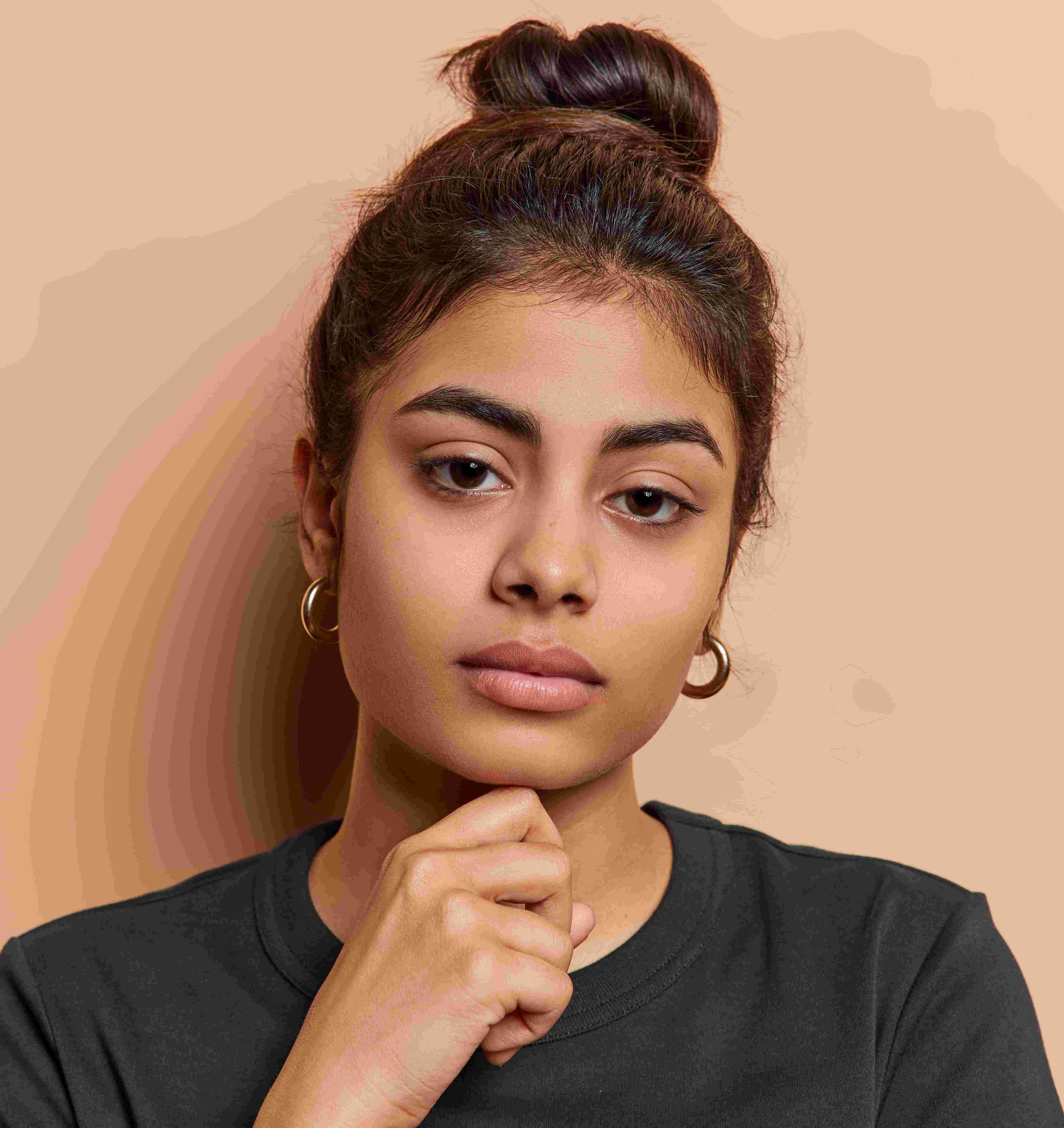 Young woman with dark hair in a bun resting her chin on her hand against a beige background.