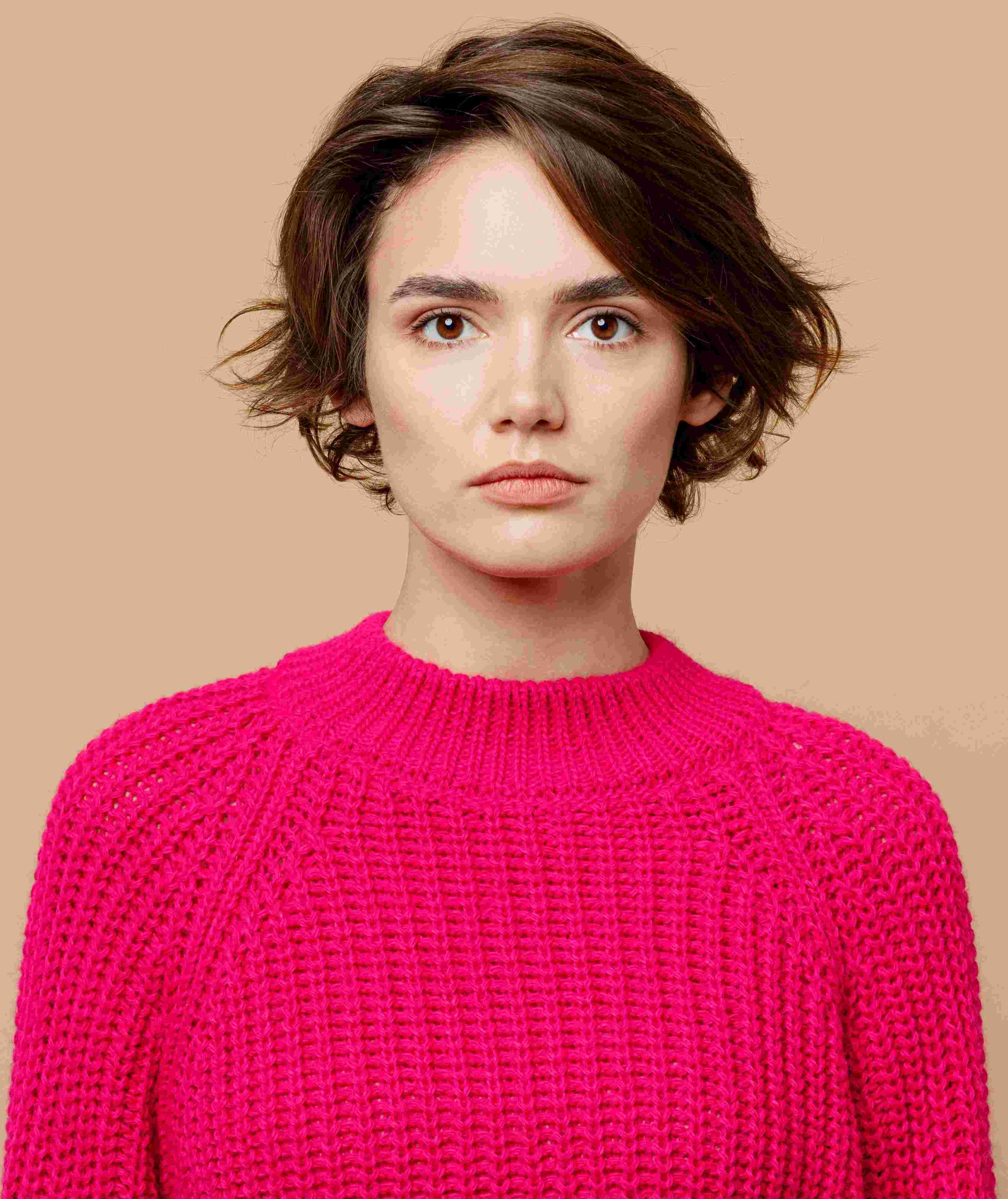 Young woman with short brown hair wearing a bright pink knit sweater against a beige background.