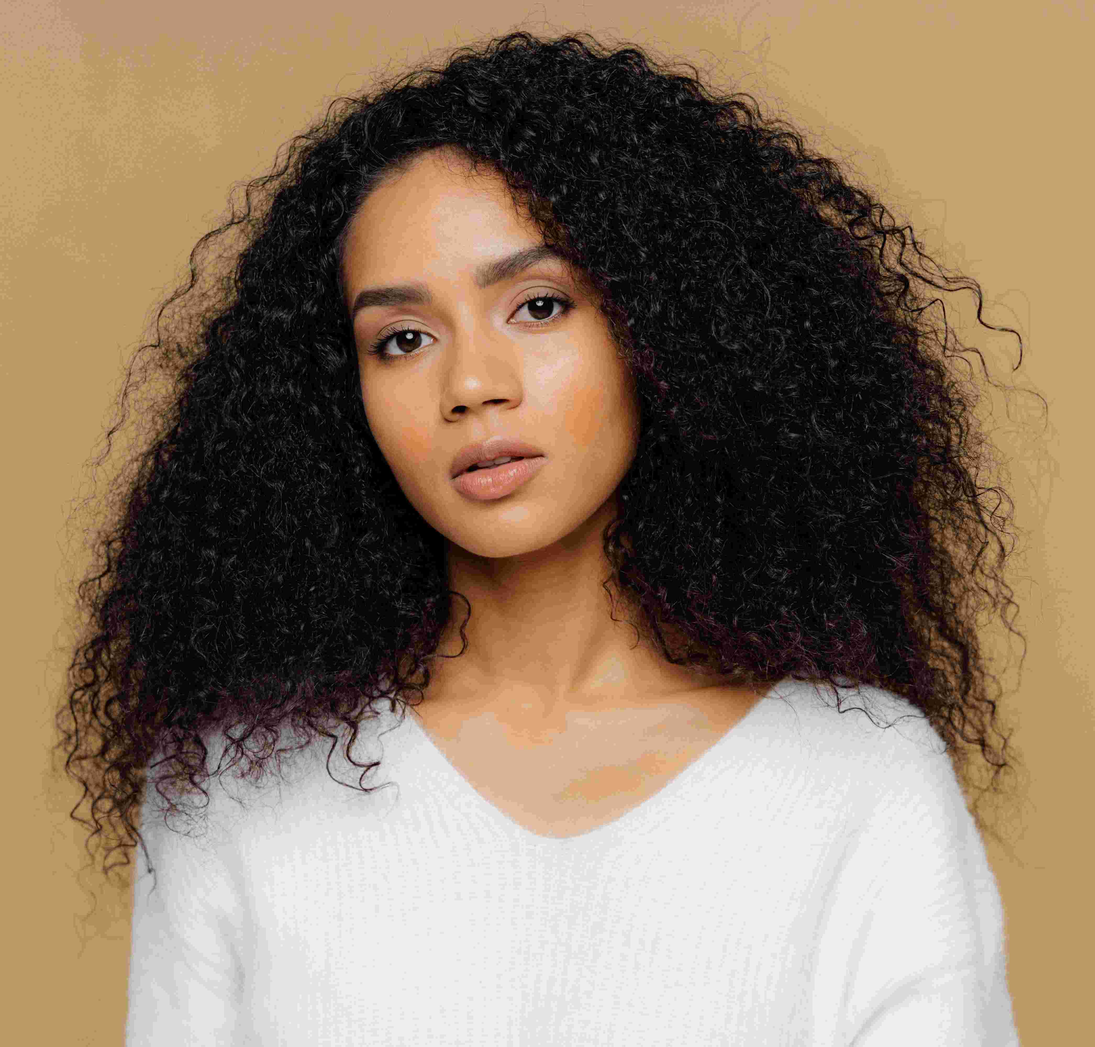 Young woman with voluminous curly black hair wearing a white sweater against a beige background.