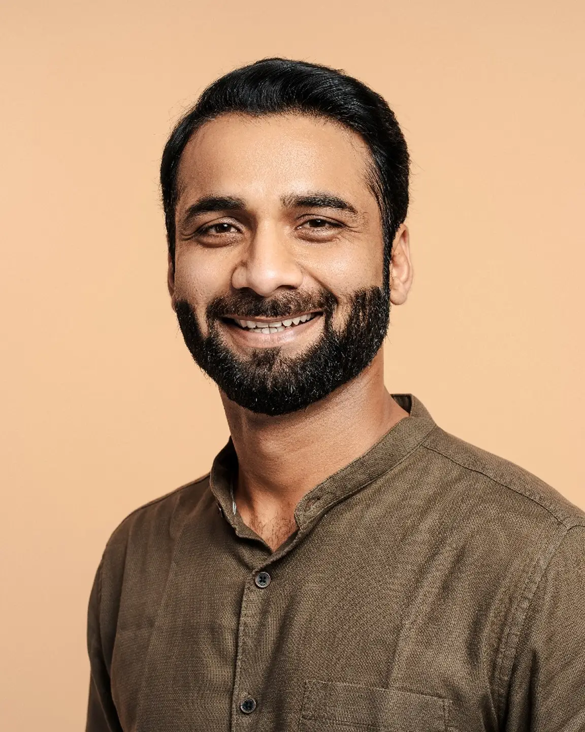 Smiling man with a beard wearing a brown buttoned shirt against a beige background.