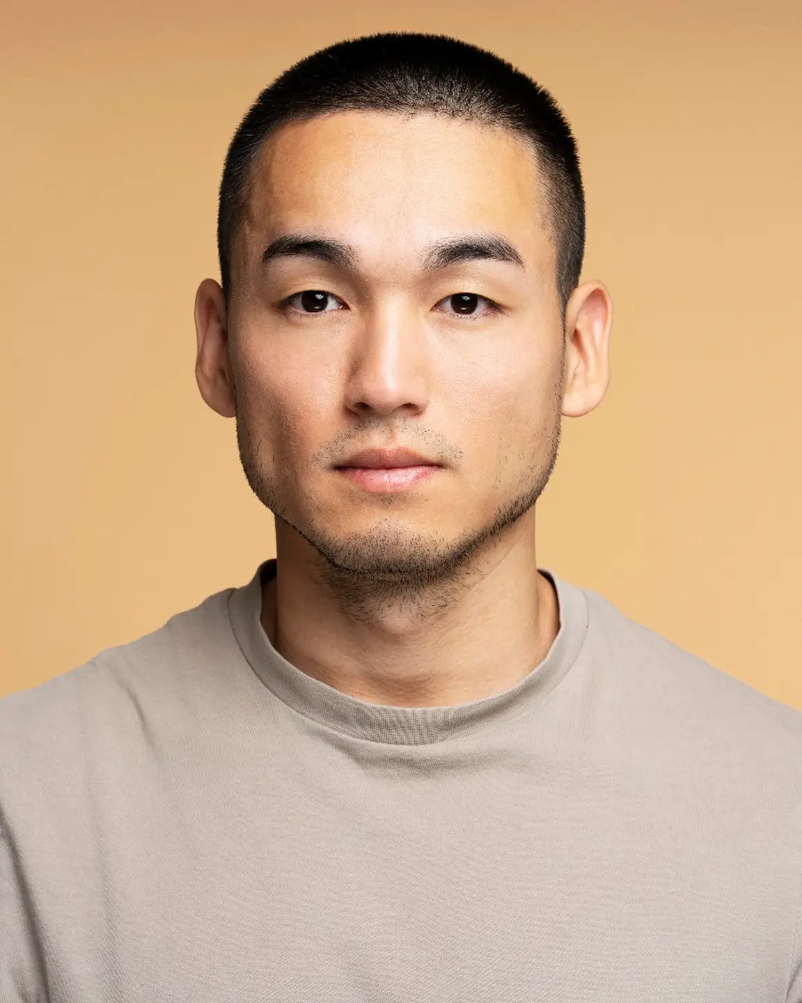 Portrait of a young Asian man with short hair wearing a light gray shirt against a beige background.