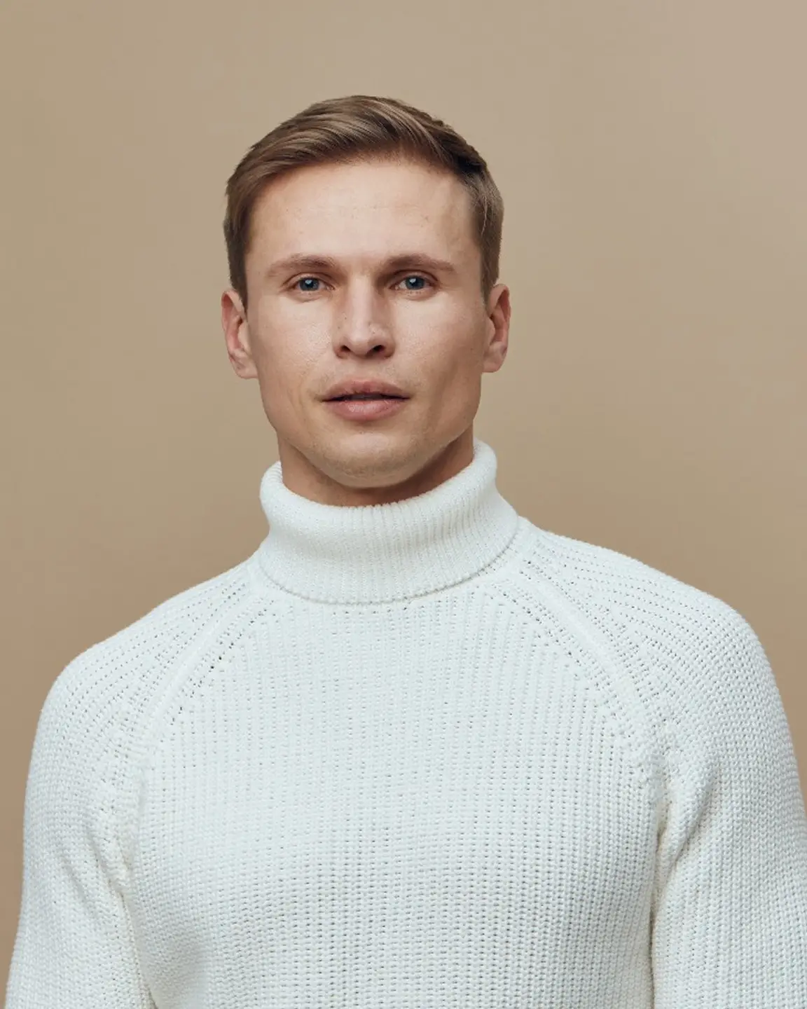 Portrait of a man with short light brown hair wearing a white knitted turtleneck sweater against a beige background.
