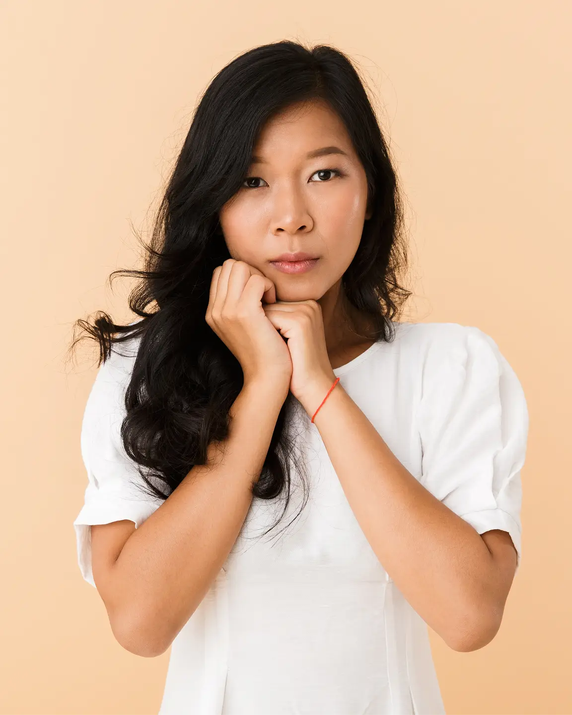 Woman with long black hair wearing a white shirt, resting her chin on clasped hands against a beige background.