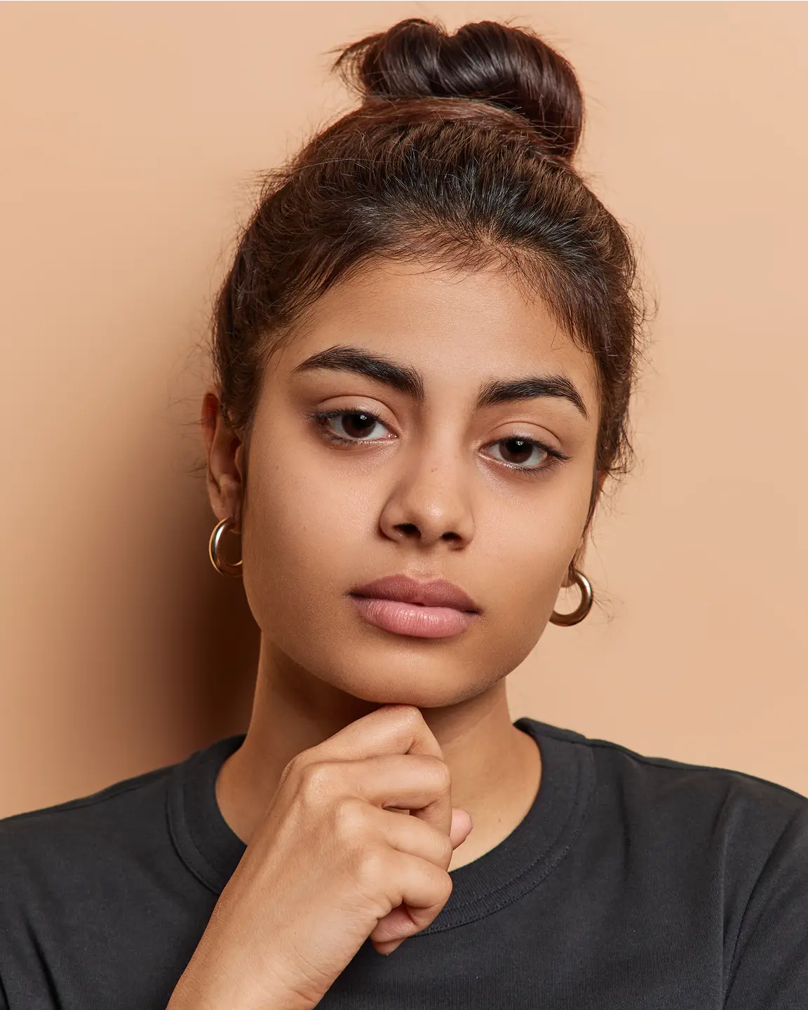 Serious young woman with dark hair in a bun, wearing gold hoop earrings and a black shirt, resting her chin on her hand against a beige background.