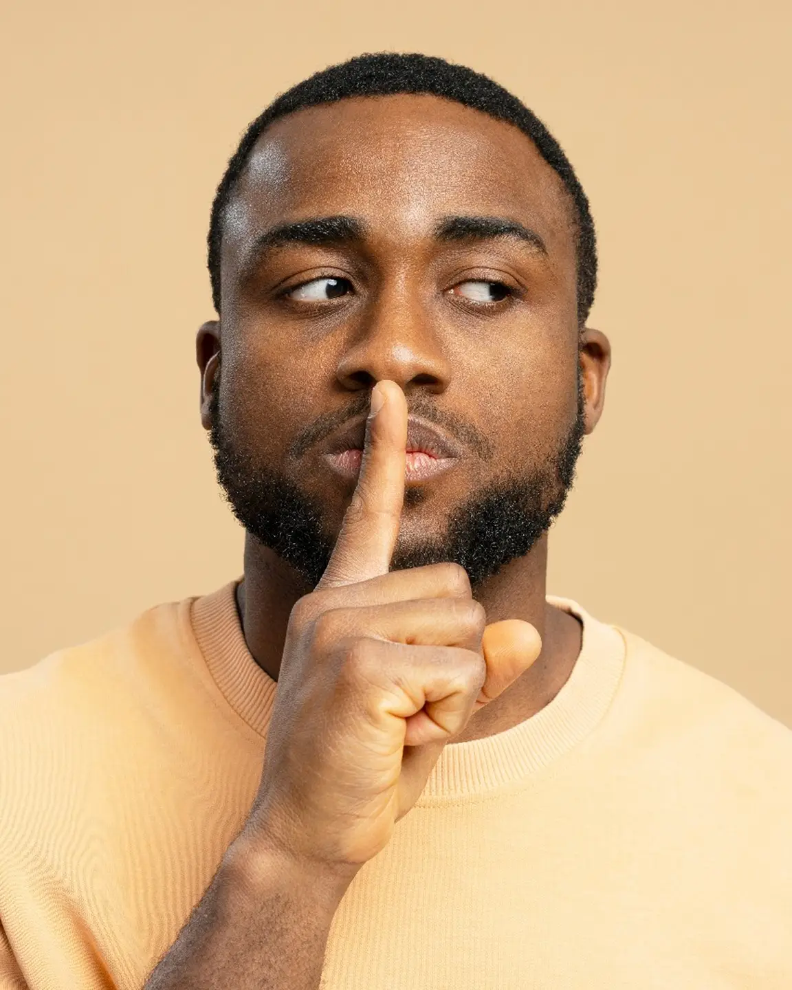 Man with a beard wearing a beige sweater holding his finger to his lips in a shushing gesture.