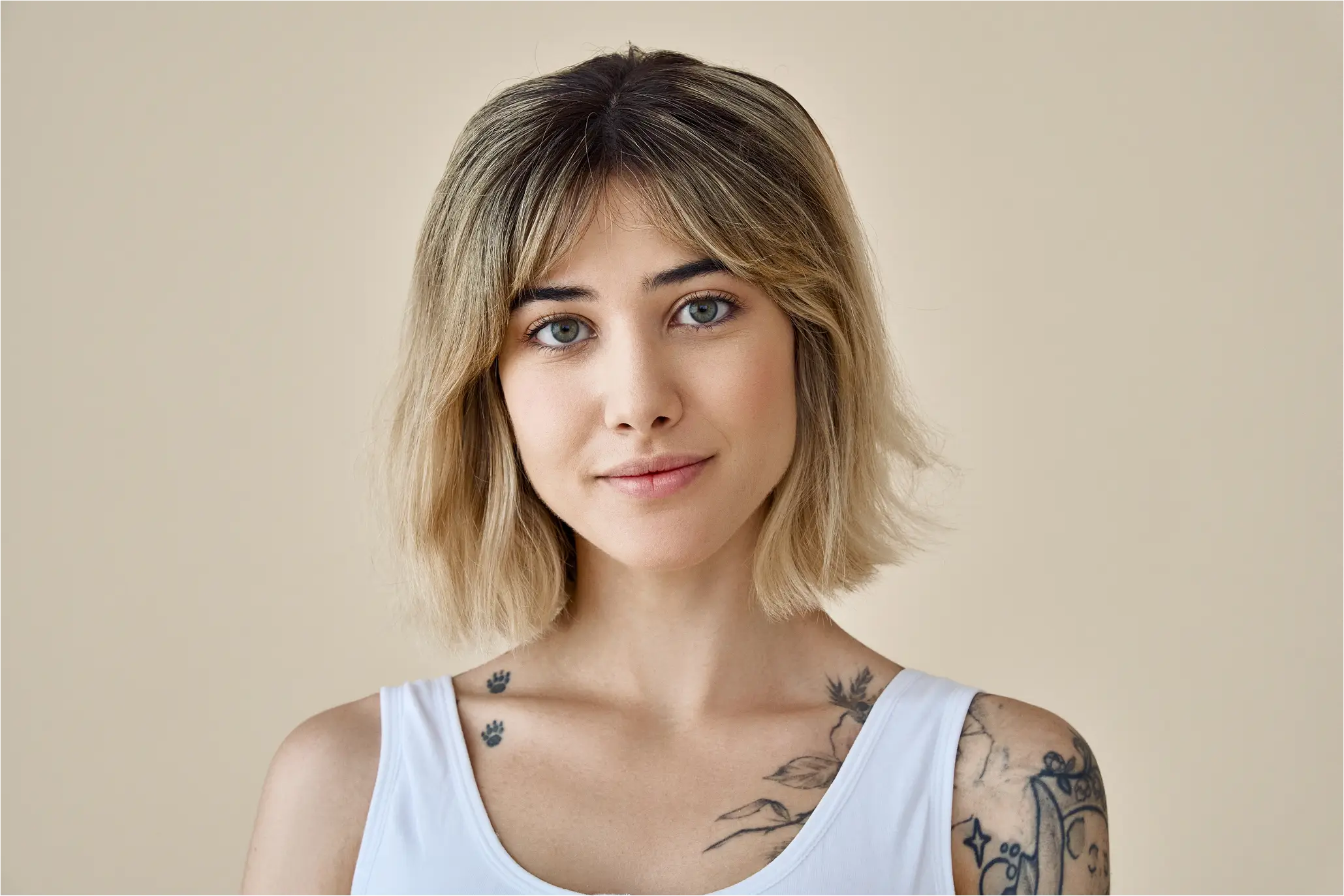 Young woman with shoulder-length blonde hair, green eyes, and tattoos on her shoulder and chest wearing a white tank top against a beige background.