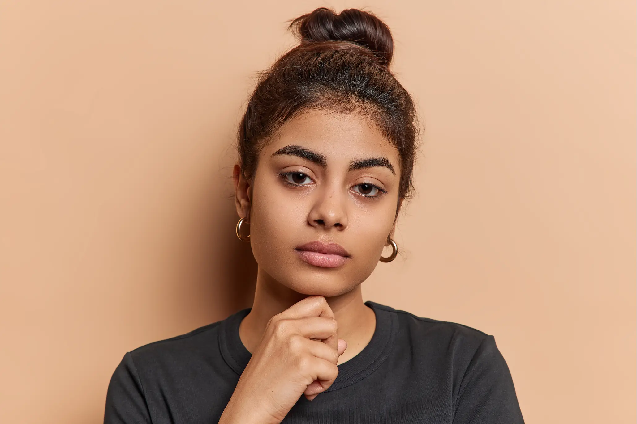 Thoughtful young woman with brown hair in a bun and hoop earrings, wearing a black shirt, resting her chin on her hand against a beige background.