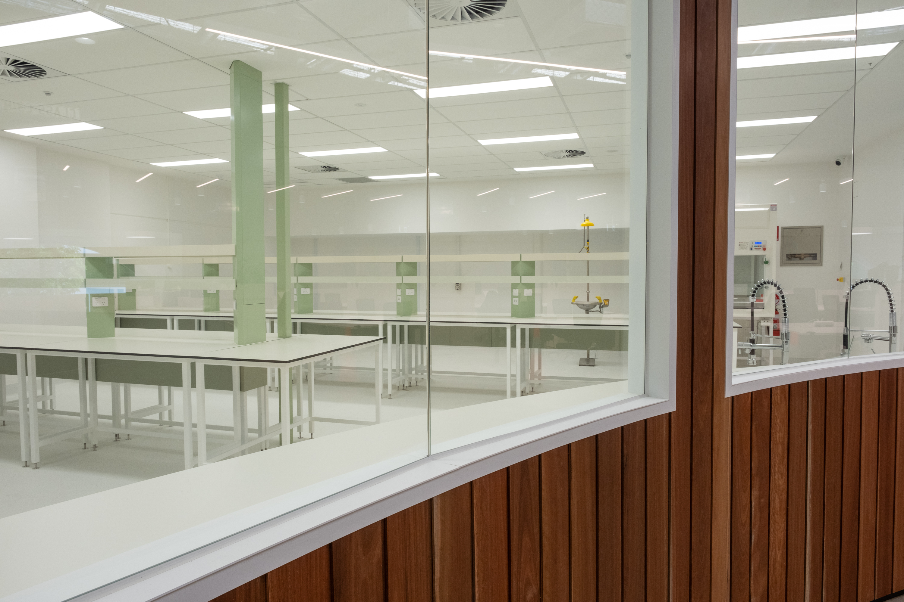Modern empty laboratory with white tables, green pillars, and a safety shower station, viewed through glass windows framed with wood panels.