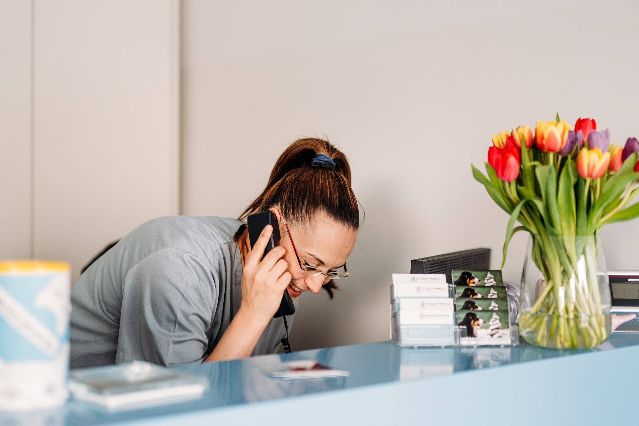 Mitarbeiterin am Empfang der Tierarztpraxis telefoniert und lächelt, daneben steht eine Vase mit bunten Tulpen.