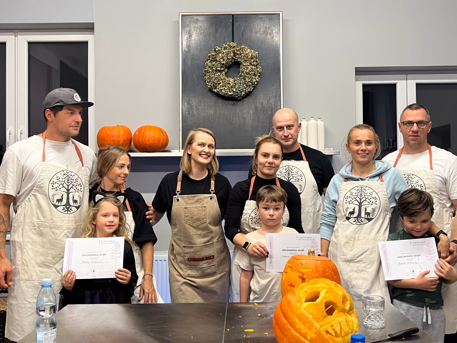 Grupa dorosłych i dzieci stojących razem w kuchni z wyciętymi dyniami na stole i certyfikatami w rękach.