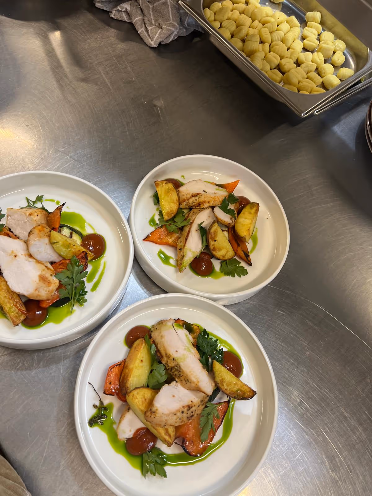Trzy talerze z grillowanym kurczakiem, pieczonymi ziemniakami, marchewką, zielonym sosem i natką pietruszki na metalowym blacie kuchennym.