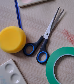 Schere, gelber Schwamm, grünes Klebeband und andere Bastelmaterialien auf einem Holztisch.