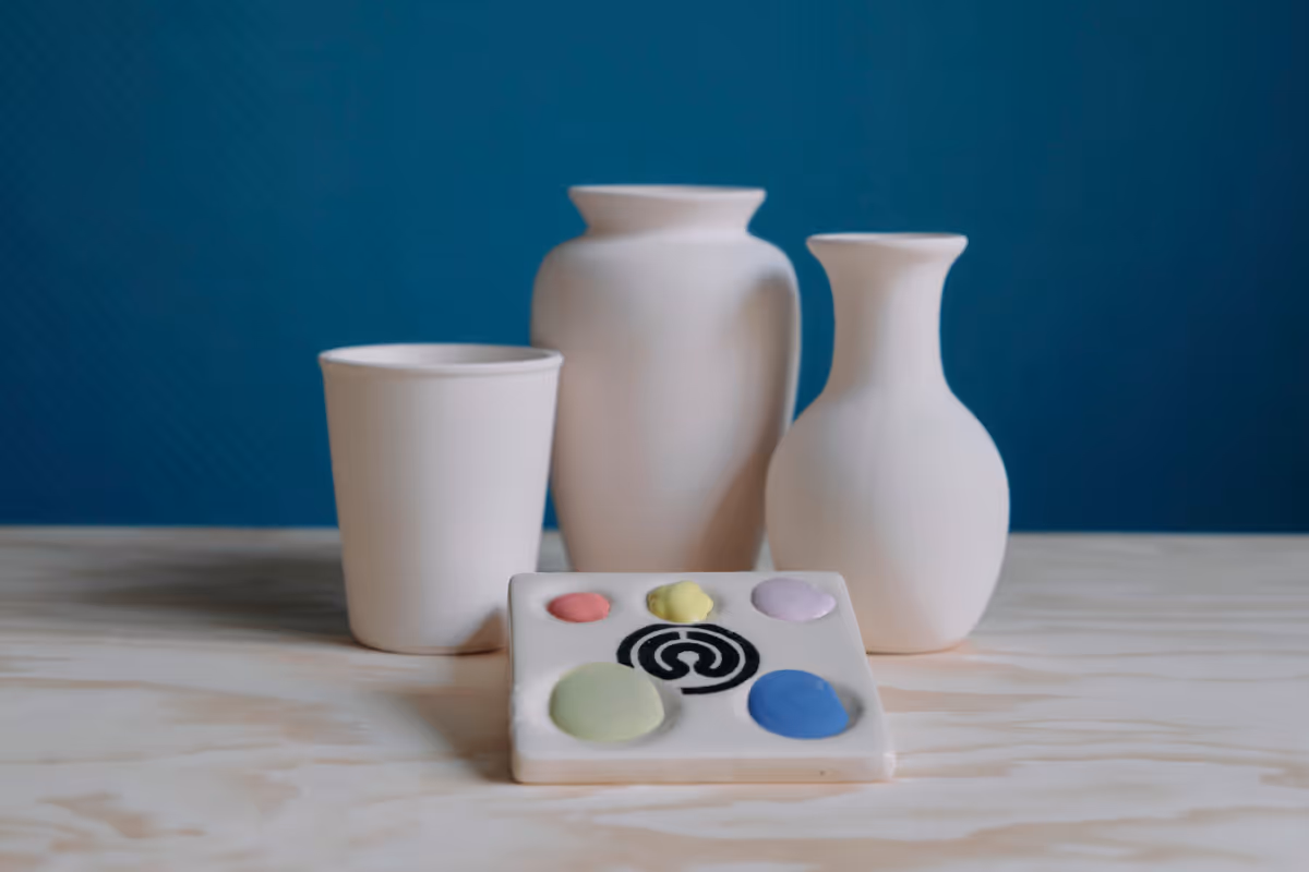 Drei weiße Keramikgefäße und eine Farbpalette mit mehreren Farbtönen auf einem hellen Holztisch vor blauem Hintergrund.