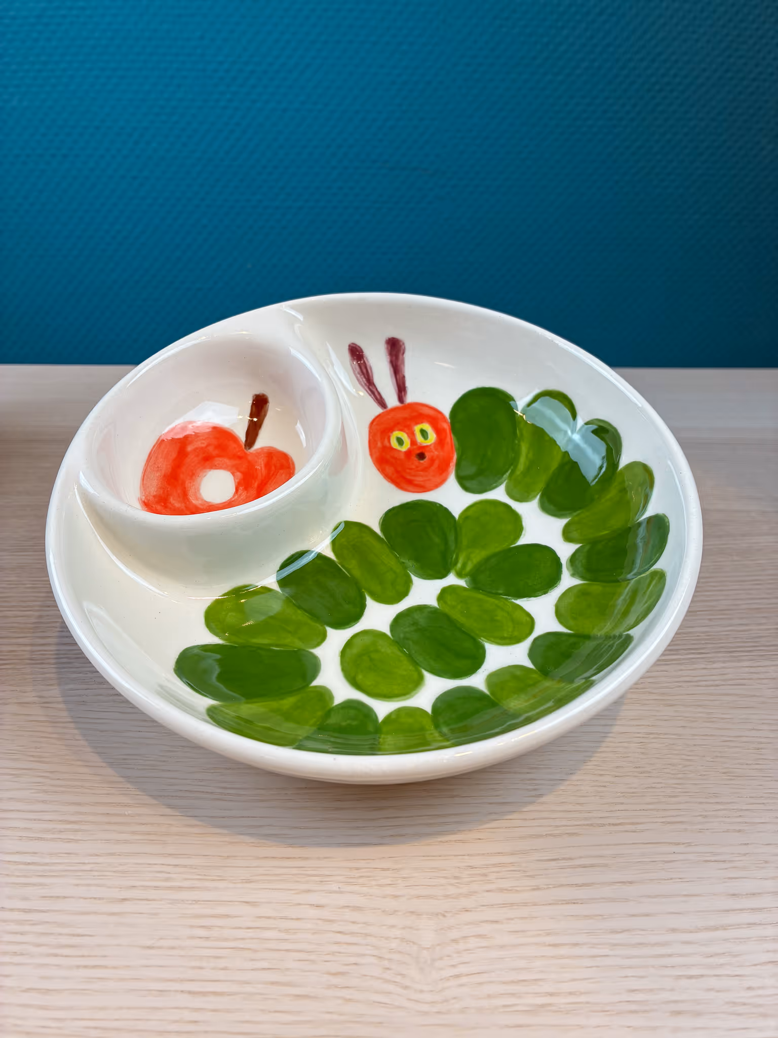 Weißer Teller mit einer Zeichnung der kleinen grünen Raupe Nimmersatt und einem roten Apfel.