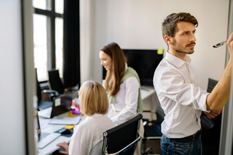A professional writes on a whiteboard while colleagues work